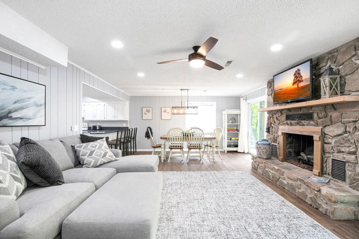 This image showcases a large gray sectional sofa facing a stone fireplace, with a dining table and chairs visible in the background. The TV is mounted on the wall, and the room is filled with natural light, giving it a cozy and inviting ambiance ideal for family gatherings and relaxing evenings.