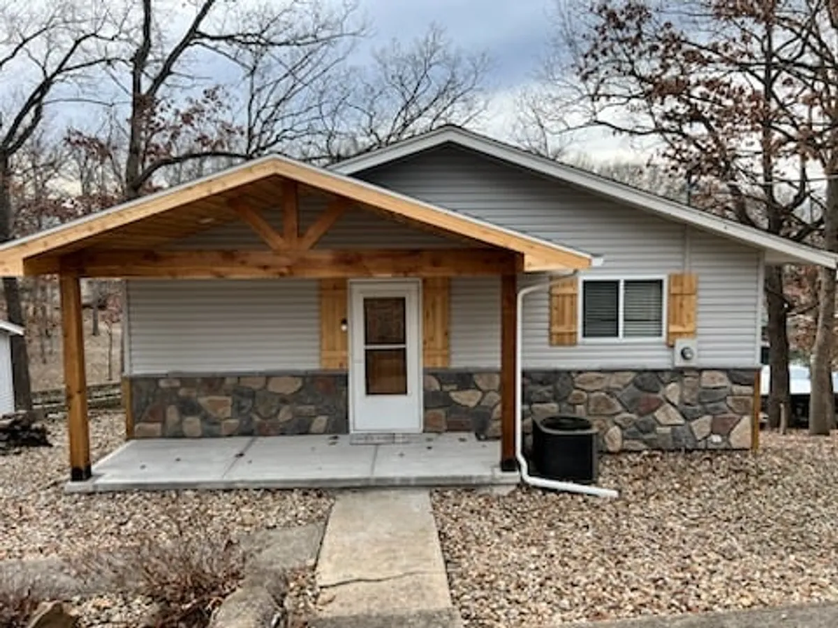 A welcoming wooden porch with a stone facade and a glass-paneled front door. Surrounded by greenery, the porch offers a cozy outdoor seating area for relaxing, socializing, or gazing at nature’s beauty.