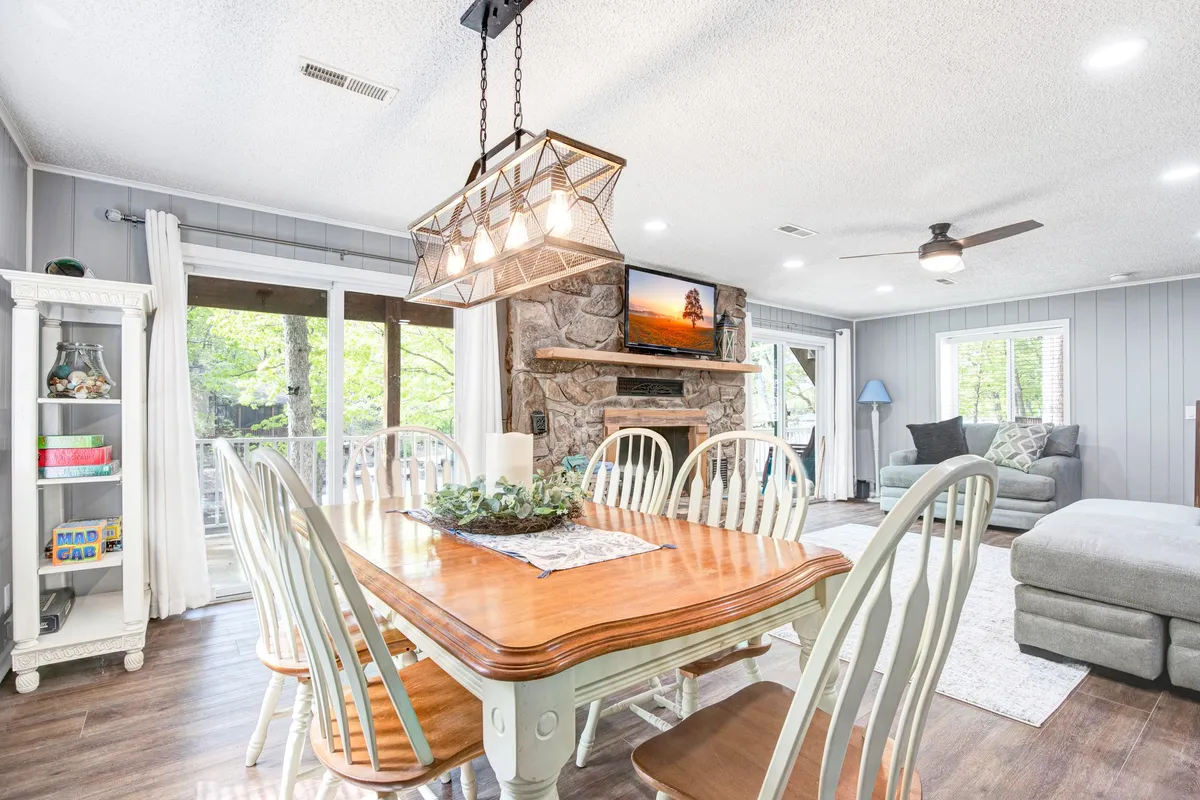 A spacious dining area with a large table set under a modern chandelier, adjacent to a fireplace and sliding glass doors. The television and comfortable seating area are visible, and the windows offer views of the surrounding nature, reinforcing the connection between the indoor social spaces and the outdoors.