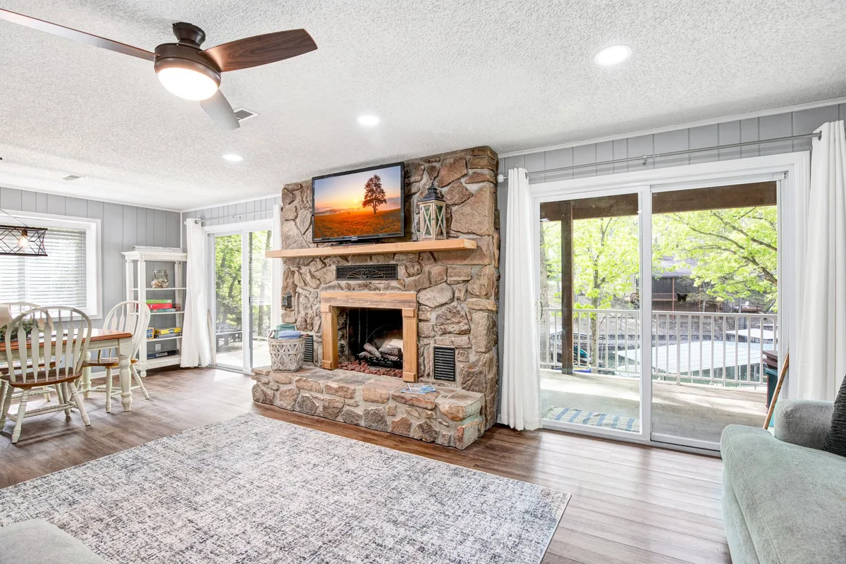 This cozy interior highlights a stone fireplace with a wooden mantel, large windows framing scenic views, and a dining area set for gatherings. The space is ideal for relaxing by the fire, sharing family meals, or enjoying board games with lake views—activities that shine during cooler months or evenings.