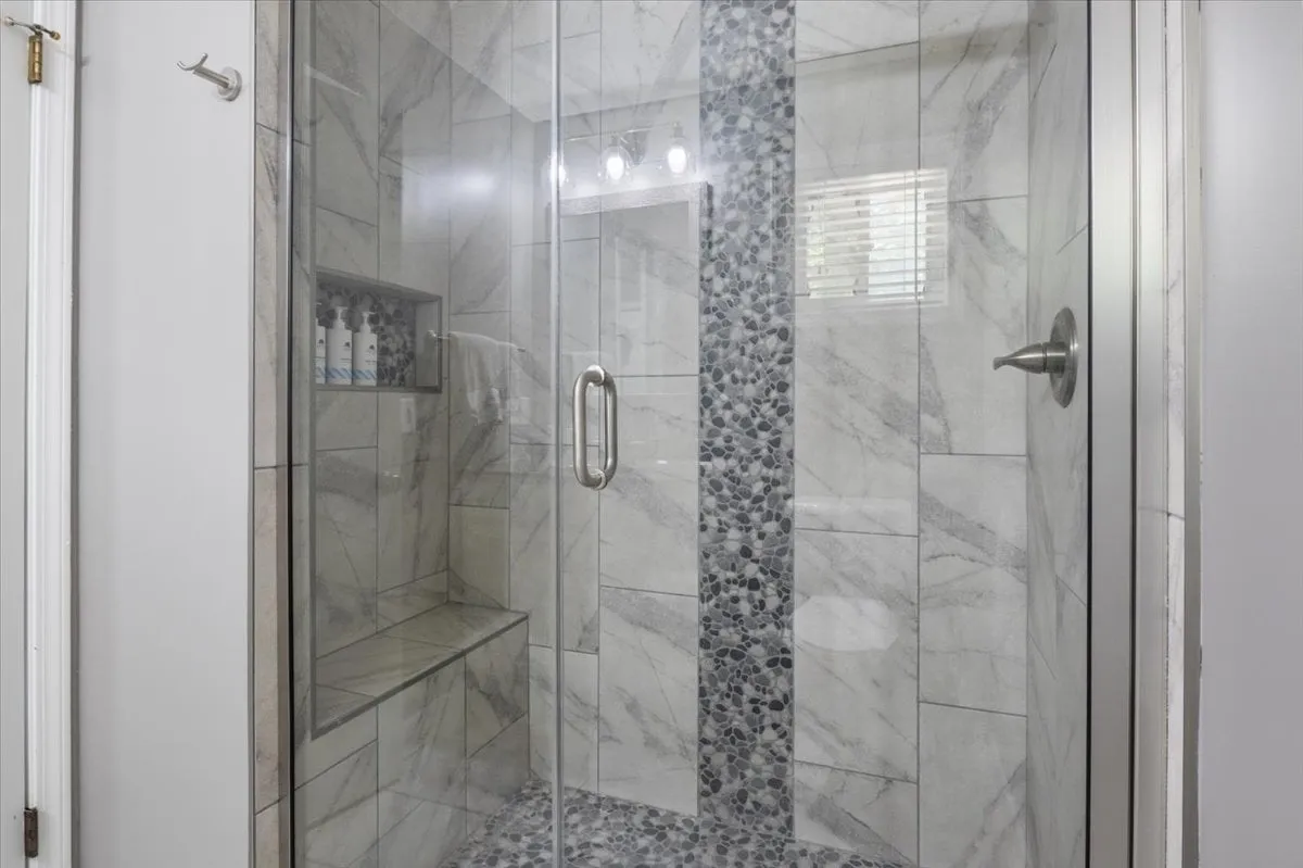 A modern bathroom with a glass shower enclosure, marble tiles, and a soothing pebble accent wall, all complemented by neatly arranged toiletries. The space feels serene, luxurious, and meticulously maintained, signaling a focus on self-care and guest convenience.