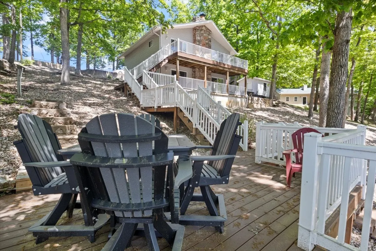 A peaceful exterior shot featuring deck chairs on a deck, the house structure in view, and stairs leading up through a forested setting. The image evokes a sense of relaxation and leisurely group moments in a natural environment.