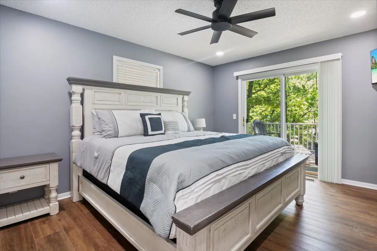 A tranquil bedroom featuring a plush bed with soft linens, a nightstand, large window, and a ceiling fan. The window offers natural light and glimpses of the lake beyond, creating a peaceful area for relaxing, reading, or waking up to sunshine. The decor is calm and inviting, emphasizing comfort and serene lake views.