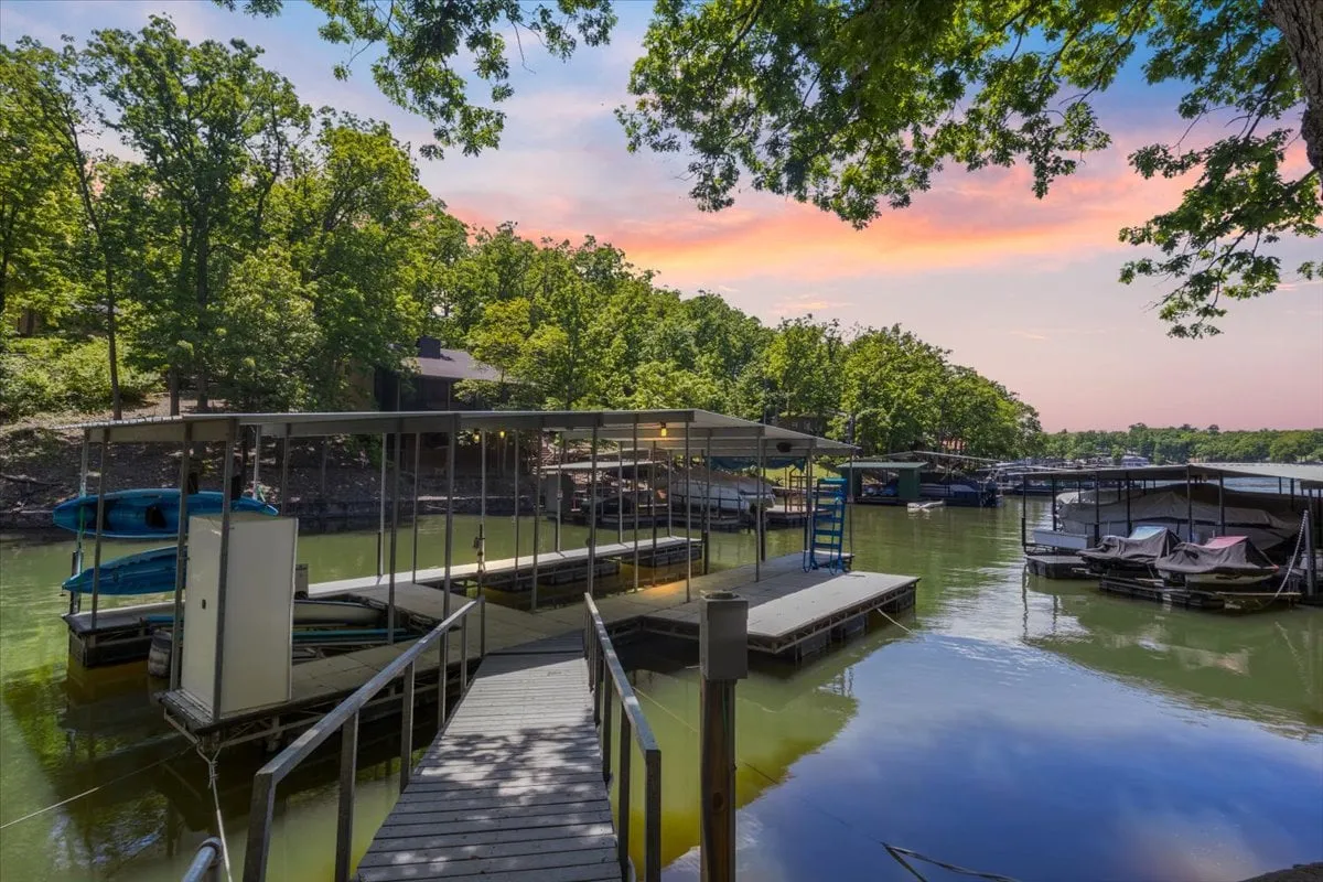 The image showcases a tranquil lakeside setting with a private dock stretching over calm water, surrounded by lush tree canopy. There are boats bobbing gently, suggesting available water activities, and the area is bathed in soft sunset light, creating a peaceful and inviting atmosphere. The spacious decks and natural beauty highlight opportunities for relaxation and outdoor fun.