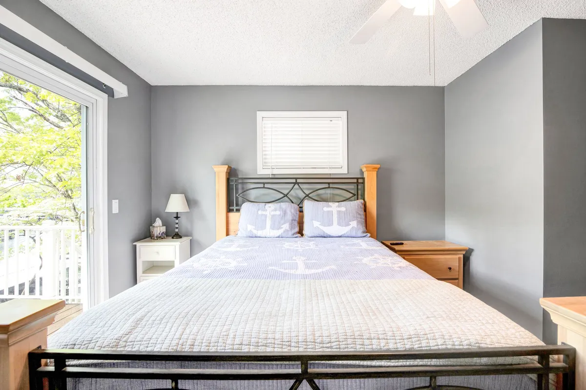 A cozy, serene bedroom featuring a bed with anchor-patterned bedding, a nightstand with a lamp, and a window offering a view of the outdoors. Natural light brightens the space, suggesting a gentle start to the day and a strong connection to the surrounding landscape.