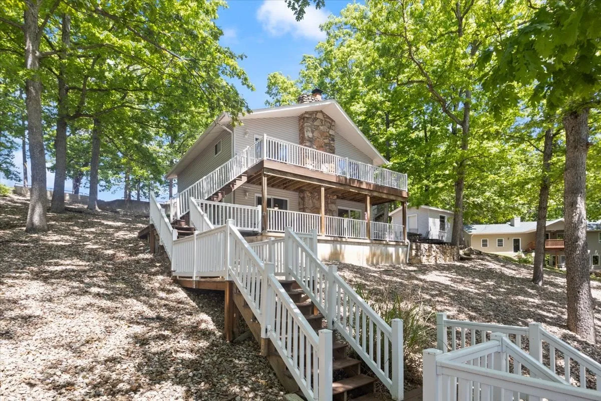 A tranquil exterior scene showing a wooden deck attached to the house, with a stone facade and stairs leading up, surrounded by tall trees. This inviting space is perfect for outdoor dining, family celebrations, and unwinding in nature.